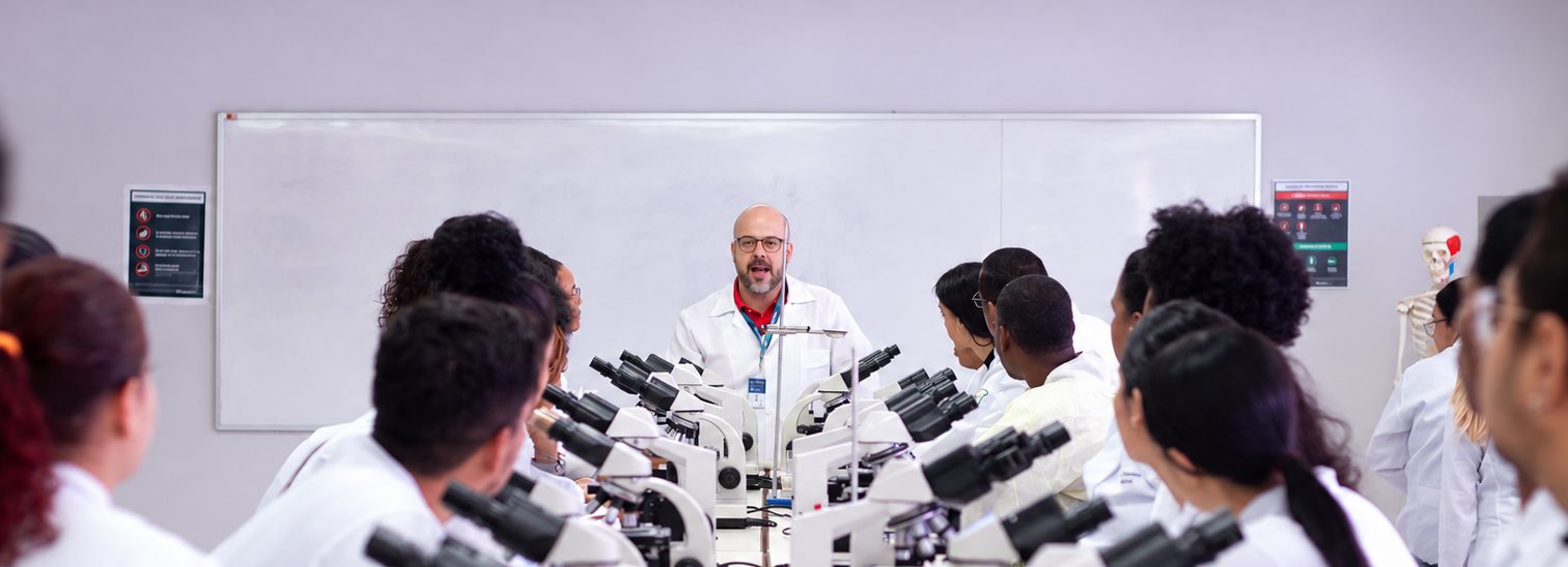 Professor dá aula em laboratório para alunos de jaleco diante de microscópios