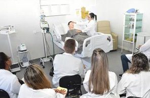 Estudantes de jaleco acompanhando uma simulação em maca hospitalar no laboratório de enfermagem