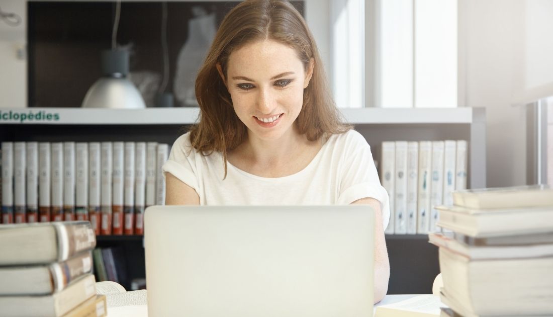 Mulher estudando no notebook com livros em volta.