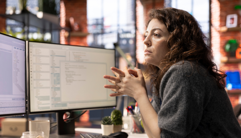 Uma mulher está parada em frente ao computador com as mãos em cima da mesa observando algumas fórmulas no computador.