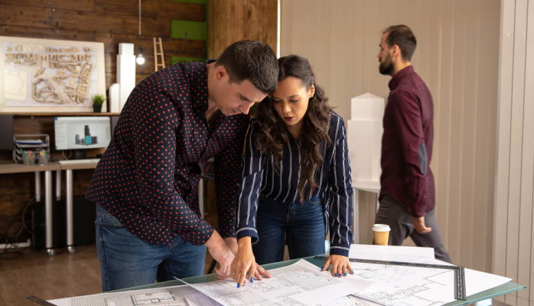 Na imagem possui uma mulher e um homem, apontando para um projeto sob uma mesa, ambos estão com as mãos apoiadas sobre o projeto, apontando para detalhes que eles percebem.
