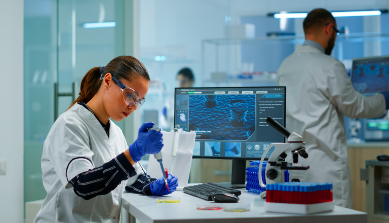Imagem de um laboratório moderno, bem iluminado, com tons claros e equipamentos científicos. Em primeiro plano, uma mulher jovem usa jaleco branco, luvas azuis e óculos de proteção. Ela está sentada ou levemente inclinada sobre uma bancada, concentrada, segurando uma pipeta e manipulando um pequeno tubo de ensaio com líquido.
