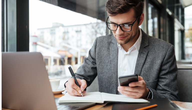 Homem segurando um celular em uma mão e na outra anotando informações em um papel. A sua frente tem um notebook aberto.