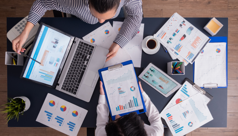 Duas pessoas analisando gráficos e relatórios em mesa com notebook, tablet e documentos.