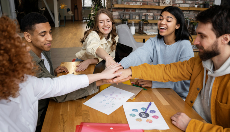 Grupo de cinco jovens sentados ao redor de uma mesa, colocando as mãos juntas no centro em gesto de união, com papéis e materiais de estudo espalhados.