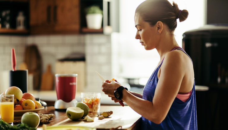 Mulher em cozinha cortando frutas frescas ao lado de um liquidificador para preparar uma vitamina.