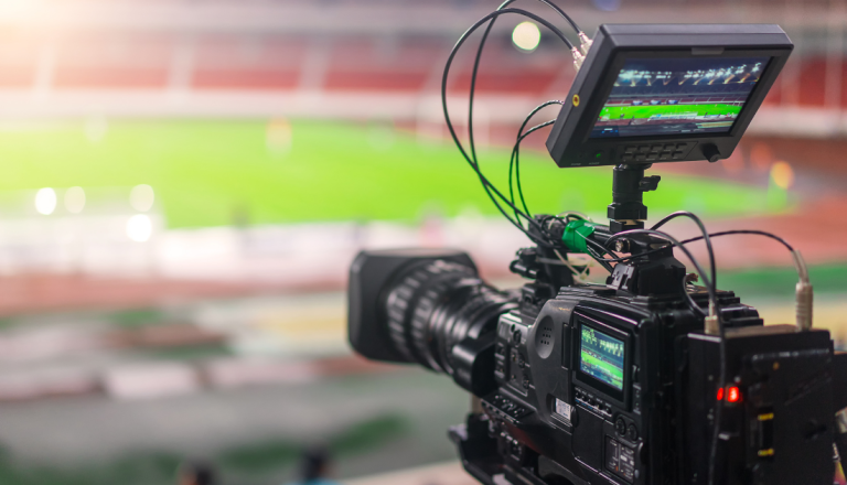 Câmera profissional de vídeo filmando um jogo de futebol em um estádio, com o campo desfocado ao fundo.