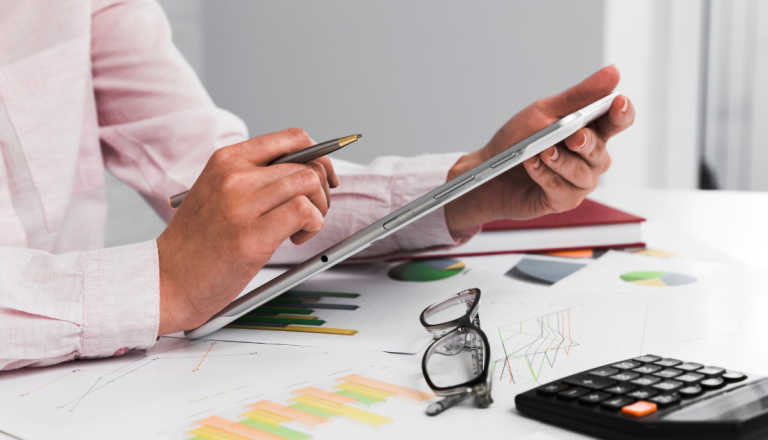 Pessoa utilizando tablet e caneta enquanto analisa gráficos financeiros sobre uma mesa com calculadora e óculos.