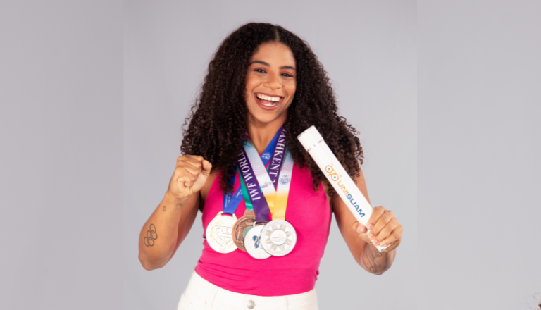Mulher sorridente com cabelos cacheados segura uma tocha e exibe três medalhas no peito, celebrando conquistas esportivas.