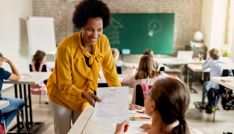Professora sorrindo entrega atividade para aluna em sala de aula enquanto outras crianças acompanham a aula ao fundo.