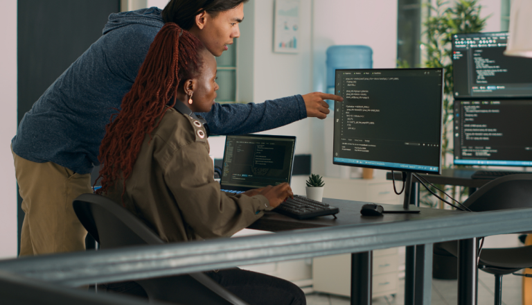 Dois estudantes de tecnologia trabalham em frente a computadores, analisando códigos em uma estação de trabalho com múltiplas telas.