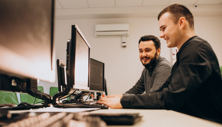 Dois homens trabalhando lado a lado em computadores de mesa em um escritório moderno, sorrindo enquanto observam as telas.
