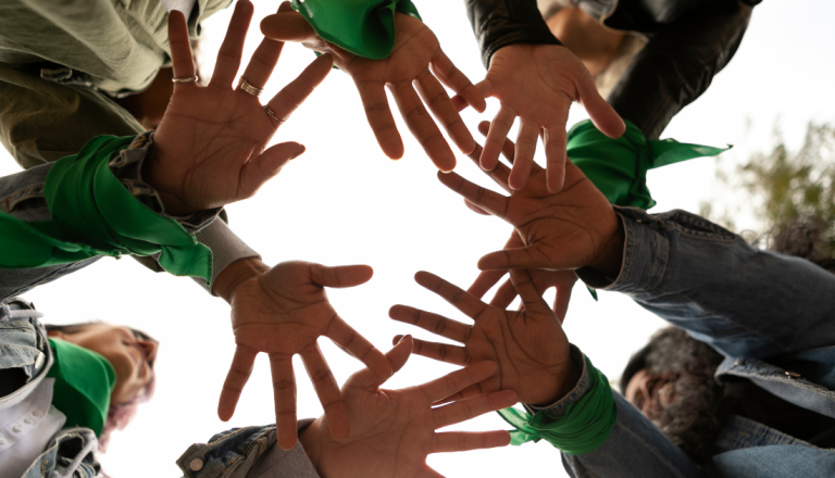 Grupo de pessoas formando um círculo com as mãos estendidas para o centro, simbolizando união, acolhimento e trabalho coletivo.