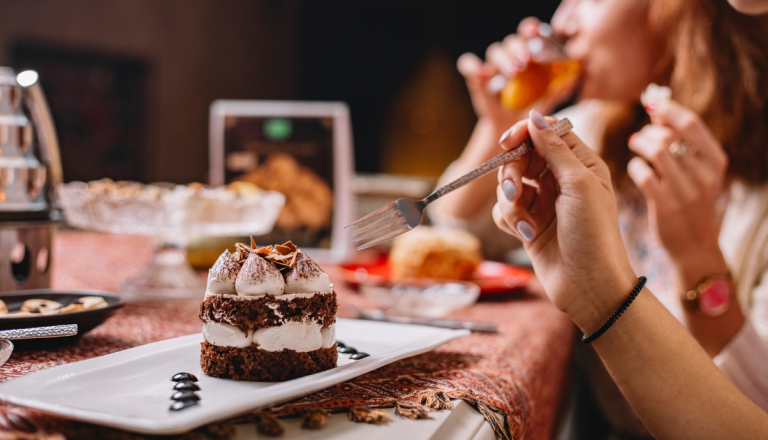 Pessoa segura um garfo diante de uma fatia de bolo de chocolate com recheio cremoso, servida em um prato, enquanto outras pessoas aparecem ao fundo em um momento de confraternização.