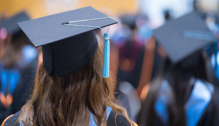 Vista de trás de formandos usando becas e capelos durante cerimônia de graduação, com foco em uma estudante em primeiro plano.