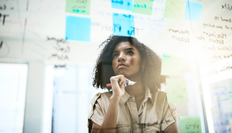 Mulher analisando anotações e post-its em painel de vidro durante momento de planejamento e desenvolvimento de ideias.