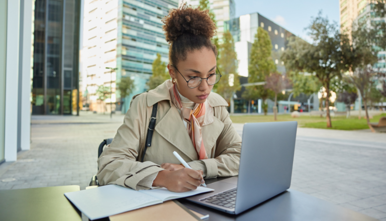 Mulher jovem estudando ao ar livre em área urbana, utilizando notebook e fazendo anotações em um caderno.