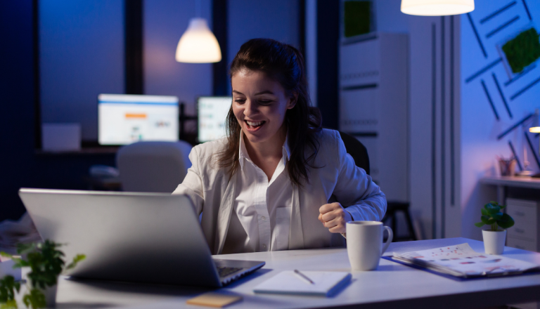 Mulher sorrindo enquanto trabalha em um laptop em um escritório à noite, com iluminação azul suave, uma caneca sobre a mesa e materiais de trabalho ao lado.