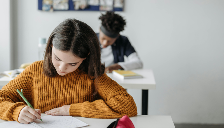 Estudante escreve em um caderno durante atividade em sala de aula, enquanto outro aluno aparece ao fundo concentrado nos estudos.