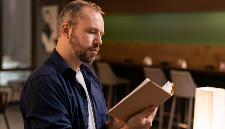 Homem sentado em ambiente interno lendo um livro, com expressão concentrada e iluminação aconchegante ao fundo.