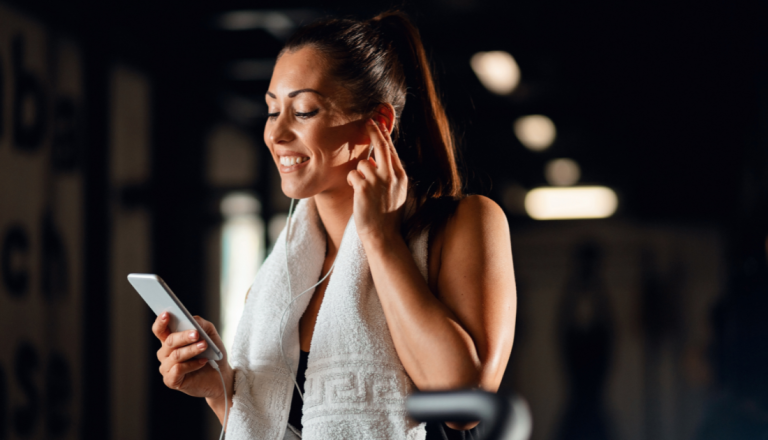 Mulher na academia olhando para o celular enquanto usa fones de ouvido, representando o consumo de conteúdos fitness e informações sobre exercícios físicos nas redes sociais.
