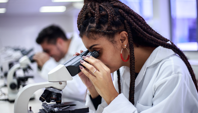 Estudante em laboratório utilizando microscópio durante atividade prática de Biomedicina, com outro aluno ao fundo.
