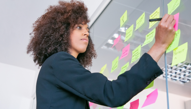 Mulher em ambiente corporativo organiza ideias em painel com post-its, representando planejamento estratégico e gestão de processos.