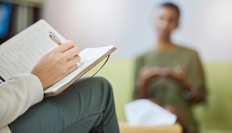Psicólogo faz anotações durante atendimento enquanto paciente conversa em sessão de terapia.