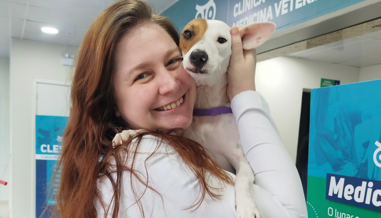 Mulher sorrindo segura um cachorro no colo dentro de clínica veterinária, destacando o cuidado e a importância da vacinação para a saúde dos pets.