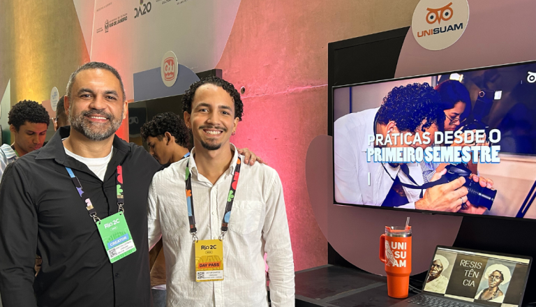 Dois participantes posam sorrindo em um estande da UNISUAM durante o Rio2C. Ao lado deles, uma tela exibe a frase “Práticas desde o primeiro semestre”, destacando experiências práticas oferecidas pela instituição.