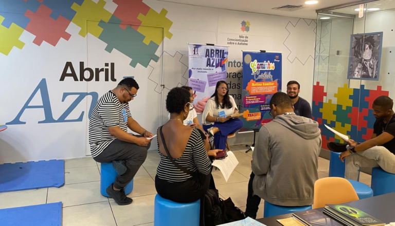 Grupo de pessoas participa de uma roda de conversa sobre conscientização do autismo em um espaço temático do Abril Azul. Os participantes estão sentados em bancos circulares, enquanto uma mediadora conduz a atividade ao lado de banners educativos sobre inclusão e rotina familiar.