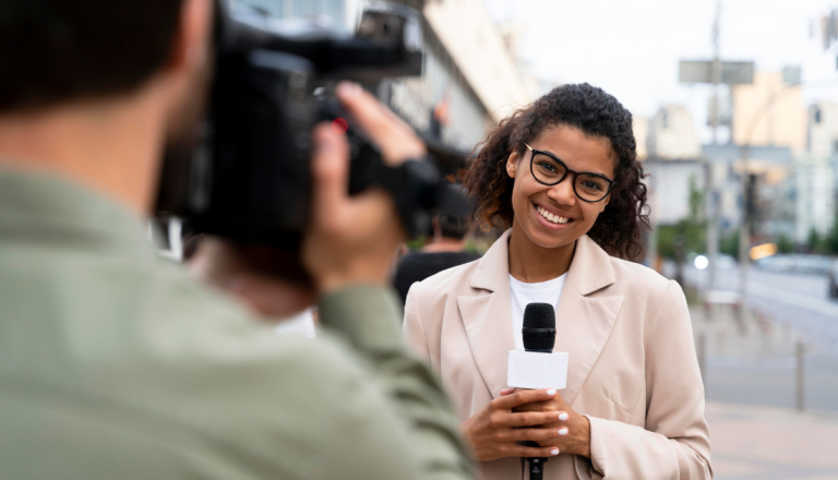 Jornalista sorri para a câmera enquanto segura um microfone durante uma reportagem em ambiente urbano, representando o compromisso com a apuração e a credibilidade da informação.
