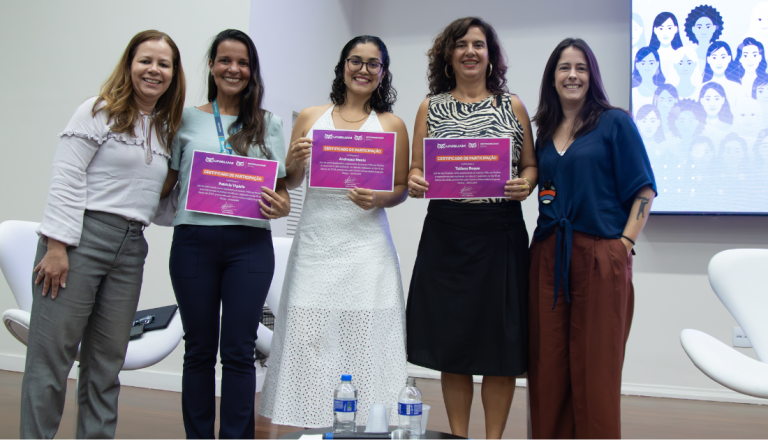 Cinco mulheres posam lado a lado durante evento da UNISUAM. Três delas seguram certificados de participação em uma atividade voltada ao protagonismo feminino.