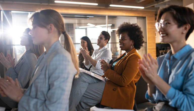 Grupo de pessoas participa de um encontro em um ambiente corporativo, sentadas e aplaudindo durante uma apresentação ou palestra, em um espaço moderno e iluminado.
