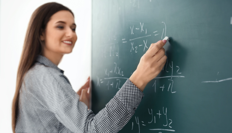 Professora sorridente escreve fórmulas matemáticas em um quadro verde com giz, durante uma aula. A imagem representa o processo de ensino, a construção do conhecimento e a atuação docente no ambiente educacional.