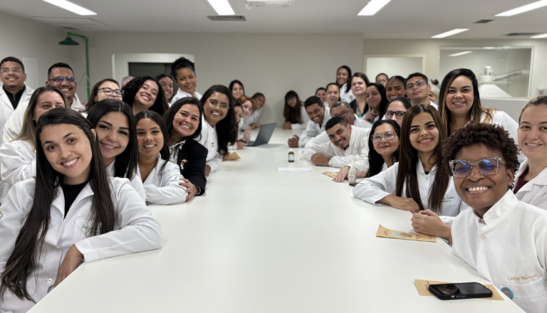 Grupo de estudantes e professores usando jalecos brancos posa ao redor de uma grande mesa em um laboratório de ensino. Todos sorriem para a câmera, em um ambiente amplo e iluminado, representando um momento de integração acadêmica.