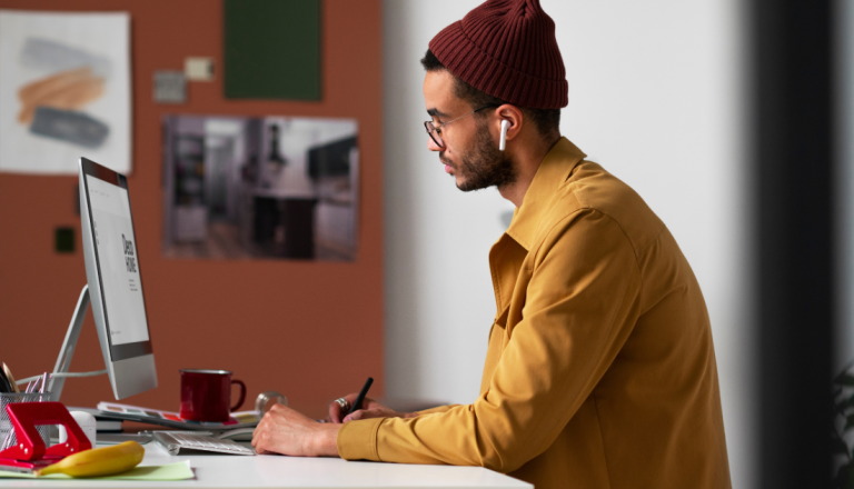 Pessoa sentada em uma mesa de trabalho usando computador de mesa, fones de ouvido sem fio e gorro, enquanto faz anotações em um ambiente interno.
