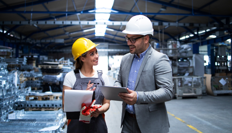 Duas pessoas utilizando capacetes de segurança conversam dentro de uma fábrica. Uma delas segura uma prancheta e a outra um tablet, enquanto analisam processos industriais em um ambiente de produção.
