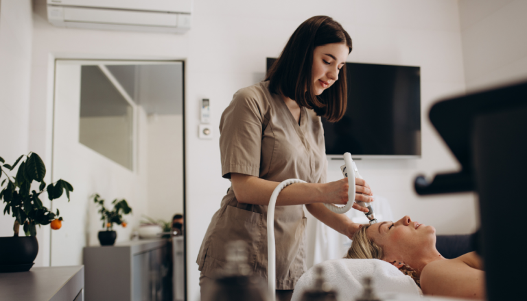 Profissional de estética realiza um procedimento facial em uma cliente deitada em uma maca, utilizando um equipamento específico para cuidados com a pele, em um ambiente moderno e acolhedor de clínica estética.