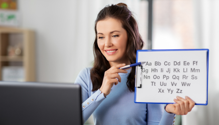 Mulher sorridente participa de um atendimento on-line e segura uma prancheta com o alfabeto em letras maiúsculas e minúsculas, apontando para as letras com uma caneta. A imagem representa a atuação do fonoaudiólogo no desenvolvimento da linguagem, da fala e da alfabetização infantil.
