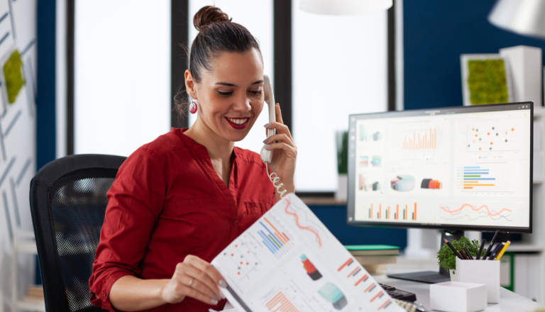Mulher sorridente em ambiente corporativo fala ao telefone enquanto analisa relatórios com gráficos impressos e acompanha indicadores exibidos em um monitor, representando o uso de dados para tomada de decisões estratégicas em vendas.