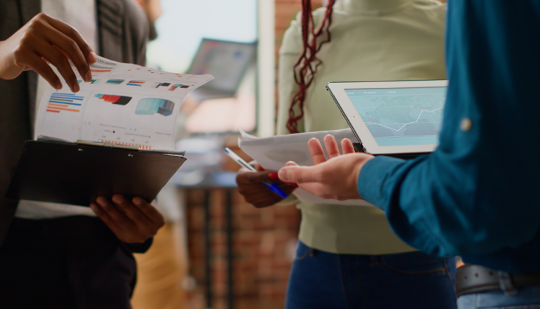 Grupo de profissionais analisa gráficos, documentos e dados em um tablet durante uma reunião, representando a tomada de decisões estratégicas e a modernização da gestão pública.