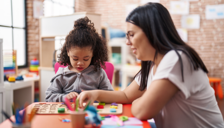 Profissional acompanha uma criança durante uma atividade lúdica com peças coloridas sobre uma mesa, em um ambiente educativo e acolhedor.
