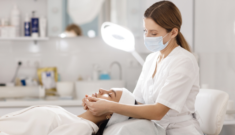 Profissional de estética realiza atendimento facial em uma cliente deitada em maca, em ambiente clínico equipado para procedimentos de cuidados com a pele.