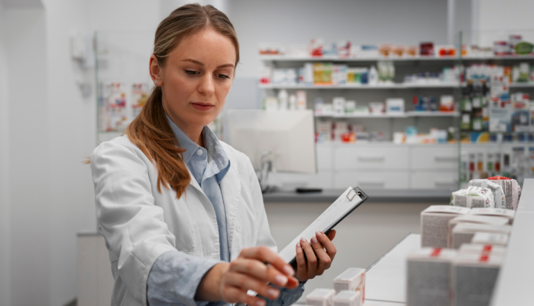 Farmacêutica de jaleco branco organiza e confere medicamentos em uma farmácia, segurando uma prancheta para realizar o controle e acompanhamento dos produtos.