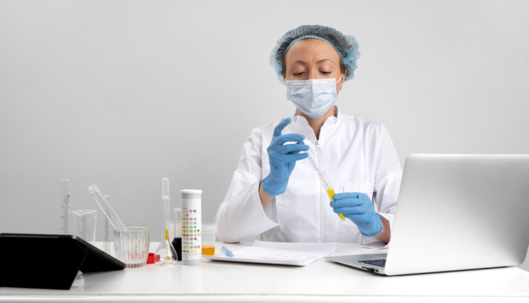 Profissional de laboratório usando máscara, touca e luvas manuseia uma pipeta e um tubo de ensaio com líquido amarelo em uma bancada com vidrarias e um notebook.