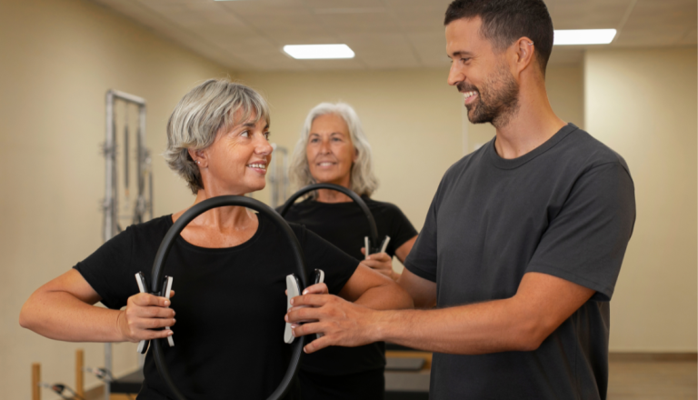 Profissional de Educação Física orienta uma mulher durante atividade física com acessórios de pilates, enquanto outra participante acompanha o exercício ao fundo.