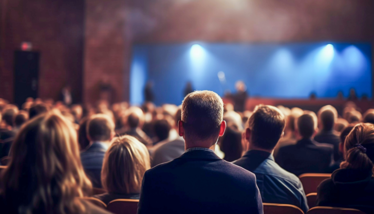Auditório lotado visto de trás da plateia. Diversas pessoas sentadas assistem a uma apresentação em um palco iluminado por luzes azuis. O foco está no público, enquanto o palco aparece desfocado ao fundo, transmitindo a ideia de evento, palestra ou conferência.