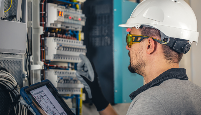 Engenheiro eletricista utilizando capacete e óculos de proteção realiza inspeção em painel elétrico, consultando um dispositivo com esquema técnico.Engenheiro eletricista utilizando capacete e óculos de proteção realiza inspeção em painel elétrico, consultando um dispositivo com esquema técnico.