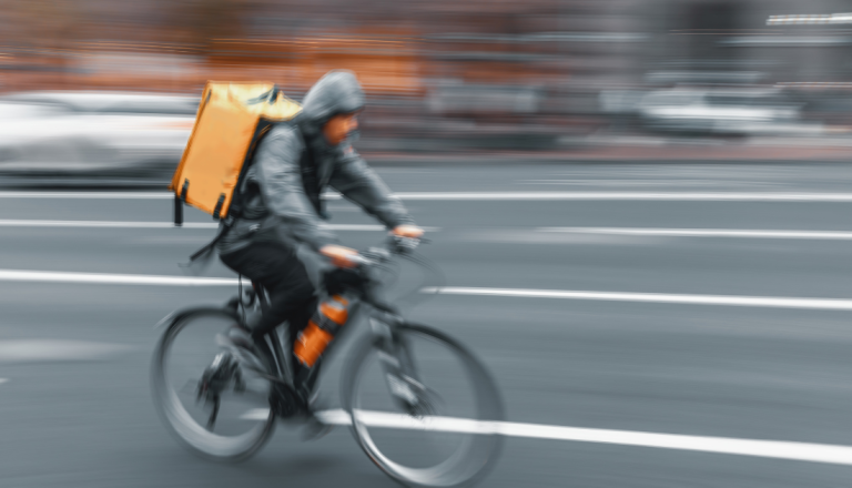 Entregador em uma bicicleta pedala por uma via urbana com mochila térmica nas costas, em uma cena com efeito de movimento que representa a agilidade das entregas rápidas.